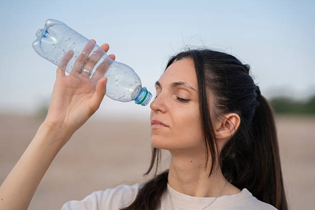 In a vast desert landscape, a woman examines an empty water bottle, expressing frustration and concern over her lack of hydration in the intense heat