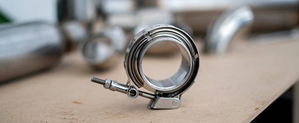 An intricate v-band car exhaust component rests on a wooden table in a workshop, surrounded by shimmering metallic parts