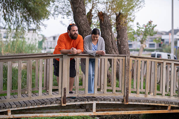 Couple Watching Scenery from Small Bridge Outdoors. Young Couple Enjoying the View from a Wooden Bridge. Young Couple Standing on Small Bridge Observing Surroundings.