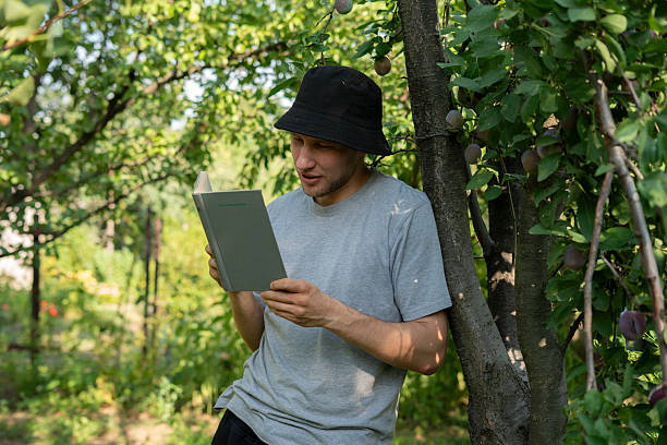 istock A person sits calmly against a tree, deeply engaged in reading a book while surrounded by lush greenery and fruit-bearing branches, enjoying a serene day outdoors. 2252916252