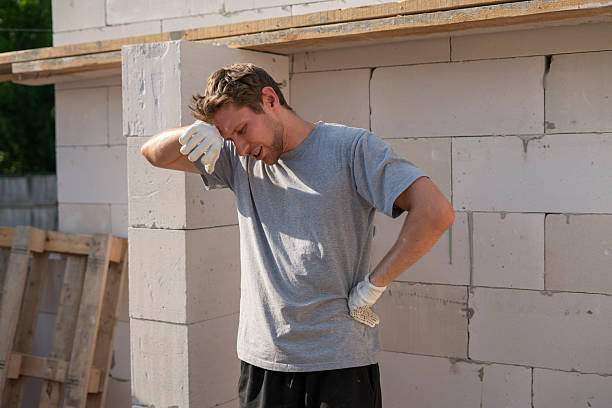 A construction worker in a gray shirt wipes sweat from his forehead while surrounded by concrete blocks on a hot day at the construction site, visibly exhausted from the labor.