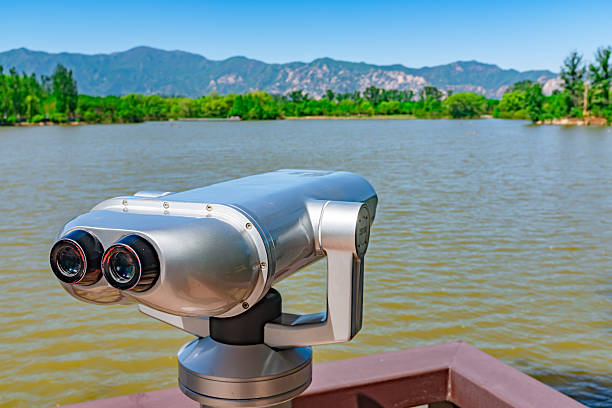 This image features a close-up of silver, metallic sightseeing binoculars mounted on a viewing deck, designed for visitors to observe waterbirds in the wetland. The device is positioned against the backdrop of a protected nature reserve, overlooking a wide expanse of calm water. The scene includes a distant shoreline thick with lush green trees and vegetation, while a rugged mountain range stands prominently against the clear blue sky in the distance, emphasizing the natural beauty of the habitat.