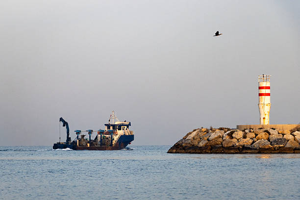 istock Dredging vessel operating near red-white striped maritime beacon on rocky breakwater—framed by calm sea, clear sky, and industrial coastal infrastructure in a serene daylight setting. 2252921749