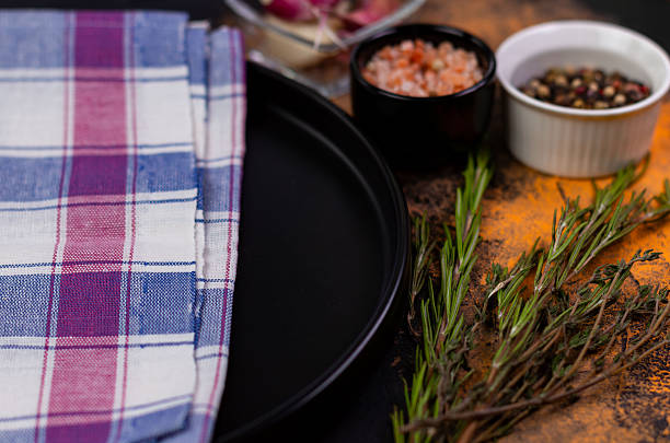A kitchen countertop displays a black plate, checked cloth, fresh herbs, and bowls of spices, all arranged for upcoming cooking activities.