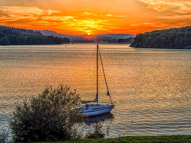 Sailboat floating on the calm lake at sunset with forest and bridge in the background