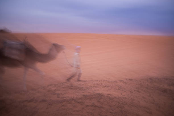 A motion-blurred camel and handler cross vast desert dunes at dusk, soft pastel light and drifting sand creating a dreamlike sense of movement and solitude.