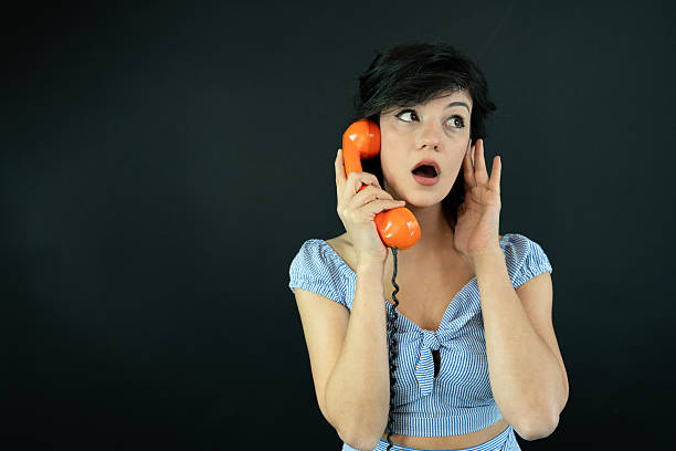 Studio portrait of a surprised young woman holding a retro orange phone. Concept of communication, unexpected news, gossip, nostalgia and emotional reaction.