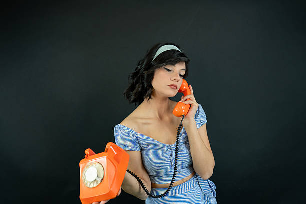 Middle aged woman calling, using red and white fixed land line telephones in vintage interior living room. Female in style dress smiling and looking at camera. Concept communication. Copy text space
