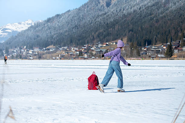 A girl in a purple sweater skates on the natural ice rink Weissensee in the Austrian Alps, warming up around a red backpack.
