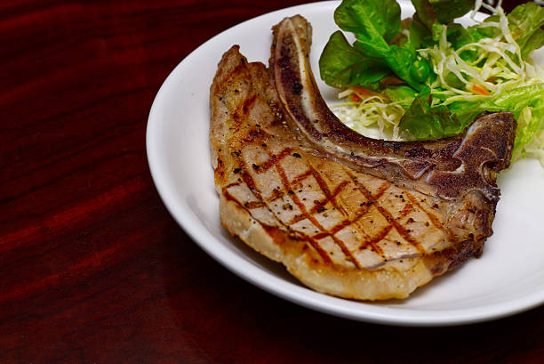 A perfectly grilled bone-in pork chop featuring distinct char marks, served alongside a fresh green side salad on a white ceramic plate over a dark wooden background