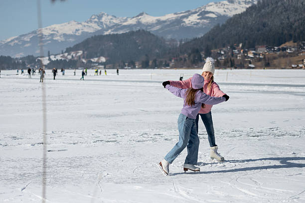A girl and her mother skates on the natural ice rink Weissensee in the Austrian Alps, warming up around a red backpack.