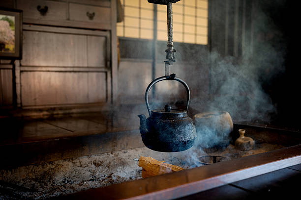 A close-up of a traditional Japanese iron kettle steaming over a burning Irori hearth with soft light in a dark room.