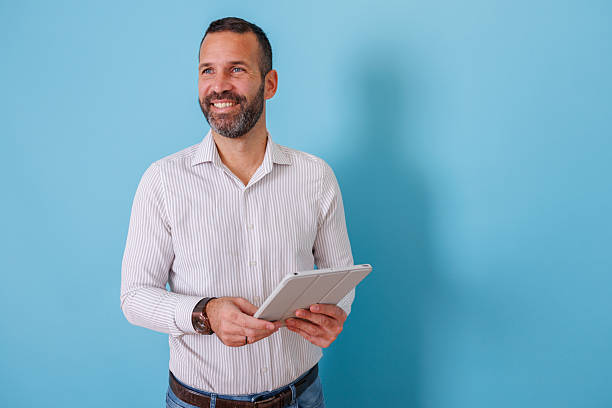 istock Confident man holding a tablet and smiling, looking away on a blue background, experiencing success and new ideas 2252984126