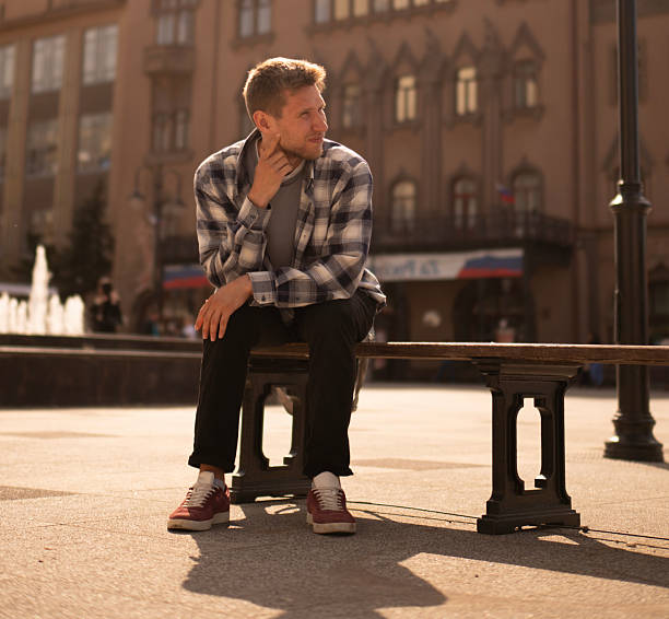 A man in a checked shirt sits thoughtfully on a bench near a striking cathedral structure as the sun sets, casting warm light