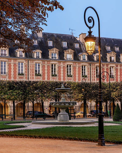 In the background, there is a grand, symmetrical building featuring red brick walls with stone dressings and steep, slate-covered mansard roofs. The roof is punctuated by elegant dormer windows.​Centered in the mid-ground is a tiered stone fountain. It sits within a circular gravel path, surrounded by well-maintained green lawns.​A prominent, ornate black street lamp stands in the foreground on the right. Its warm, golden glow suggests the photo was taken during \
