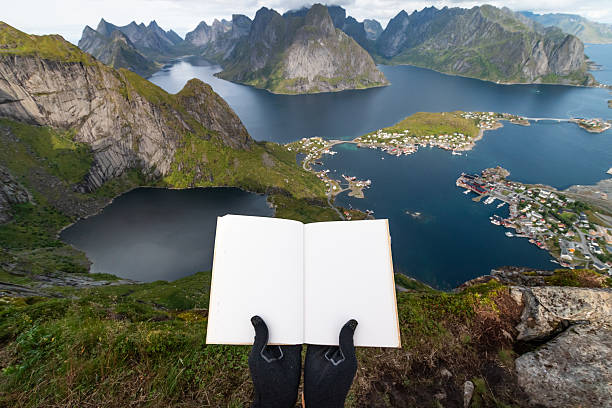 A woman standing on the Reinebringen mountain top, holding a blank notebook and looking at the Lofoten Islands and the Reine village in Norway during a sunny day of summer.