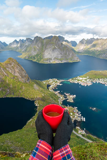 A woman standing on the Reinebringen mountain top, holding a red cup of coffee and looking at the Lofoten Islands and the Reine village in Norway during a sunny day of summer.