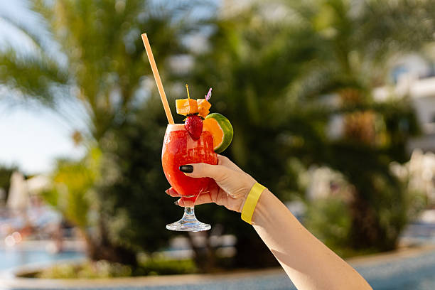 istock A hand holds up a glass filled with a bright red drink topped with fruit while enjoying time by a swimming pool. The sun shines in the background, creating a warm atmosphere 2253019365