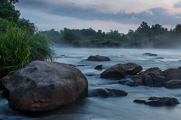 A unique view of a riverside hot spring where steaming rocks line the water's edge, set against a lush mountainous backdrop under a cloudy sky.