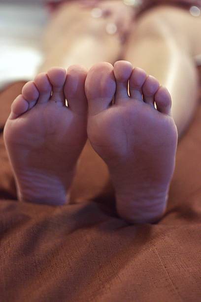 An intimate and detailed shot of the soles of a young woman's feet, capturing the texture of the skin and the natural lines of the soles. The lighting is soft and warm, creating an atmosphere of relaxation, self-care, and well-being. The blurred, earth-toned background complements the warmth of the skin, making this image ideal for health, podiatry, spa, beauty, or home lifestyle themes.
