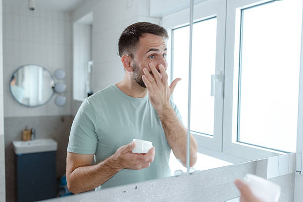 istock Man in bathroom performing morning routine, looking at mirror and applying face care 2253034296