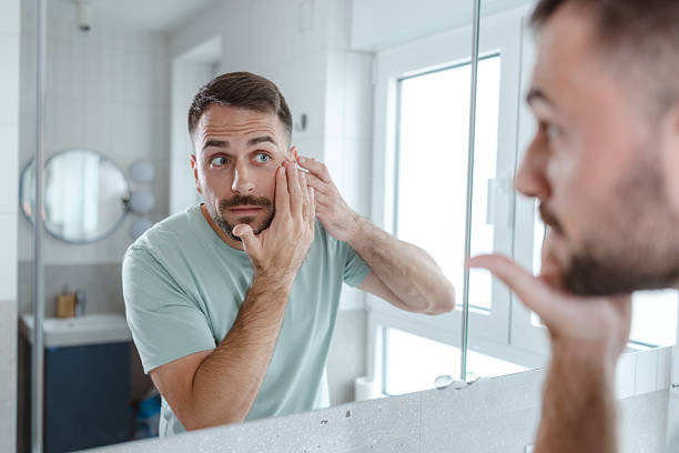 istock Man in bathroom touching his face and looking in mirror 2253034306