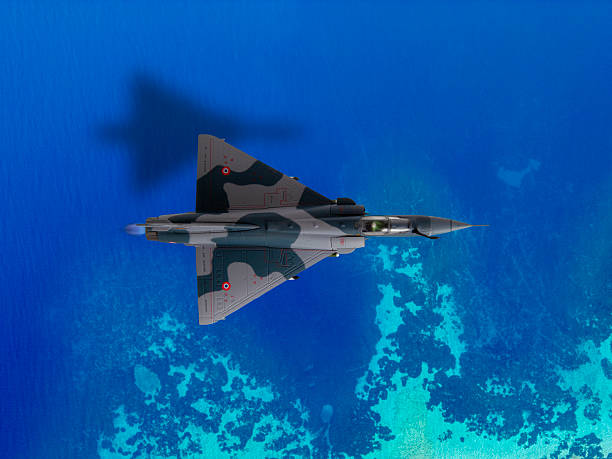An aerial view captures a camouflage-painted fighter jet soaring above a tropical sea, revealing submerged coral formations and the aircraft's shadow