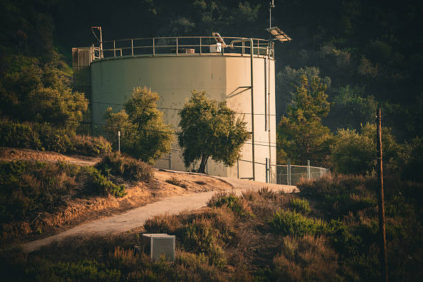 A tall, cylindrical water storage tank stands prominently on a sun-drenched, arid hillside surrounded by dense vegetation and a dirt road leading towards it