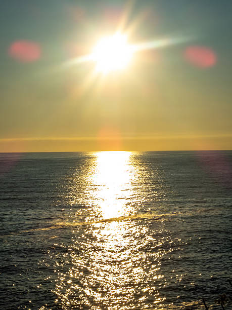 Scenic vertical shot of a bright sun setting over the sea. Golden hour light creates a sparkling reflection on the water with soft flares in Huatulco, Oaxaca, Mexico.