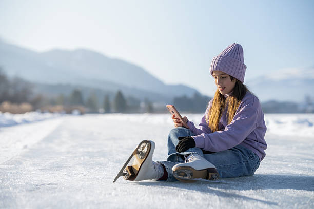 A cute teenage girl leisurely figure skating outdoors on a pond ice rink on a cold winter day. She wears a gray jacket, skirt, tights, red neck scarf and mittens for warmth as she extends arms and glides on one foot in a Lake Harriet urban park, Minneapolis, Minnesota.