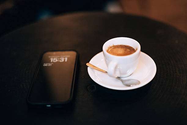 Cup of espresso or cafe crema on a white saucer with spoon placed next to a smartphone displaying time on a dark wooden café table.