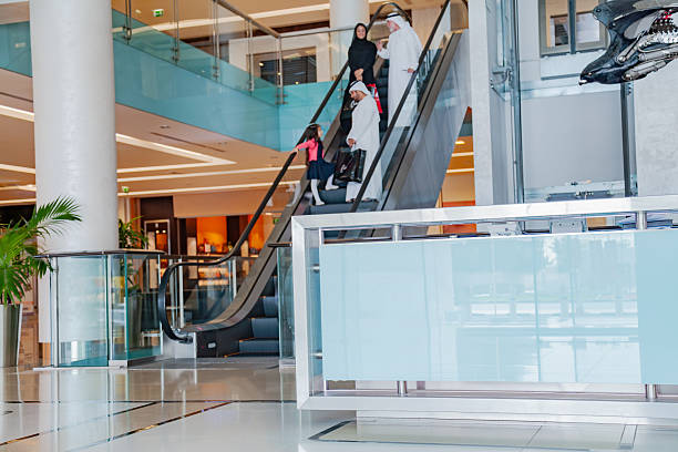 A Middle Eastern three generation family consisting of an elderly grandfather, adult parents, and a young girl walk together inside a modern shopping mall while holding hands, expressing family bonding, care, and togetherness. The grandfather wears traditional white kandura and headscarf while talking on a smartphone, the mother wears a black abaya and carries shopping bags, and the little girl wears a navy dress with a pink cardigan. The scene is captured in a bright contemporary mall corridor with glass railings, indoor plants, polished floor reflections, and a cafe and patisserie in the background, representing modern lifestyle, family values, retail experience, and everyday life in the Middle East