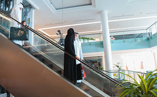 An Arab three generation family wearing traditional clothing is riding an escalator inside a modern shopping mall. The scene shows grandparents, parents, and a young daughter together, interacting and talking while moving between floors in a bright, contemporary retail center. The image is photographed from a wide angle, emphasizing glass railings, indoor plants, architectural lines, and spacious mall design, creating a lifestyle, leisure, and shopping concept with visible copy space
