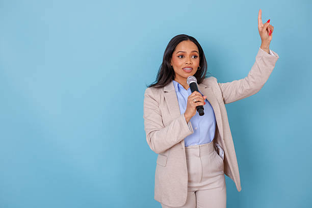 istock Businesswoman speaking into microphone, giving presentation, pointing finger up while communicating ideas 2253058717