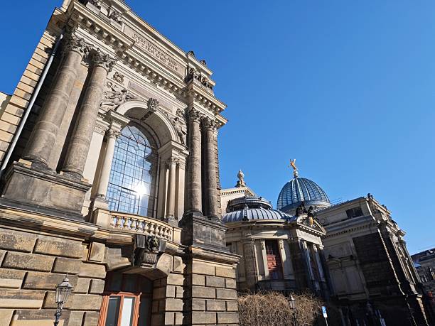 Exterior view of the Semper Opera House in Dresden captured from a low-angle perspective under a clear blue sky. The historic opera building features an ornate sandstone façade in Neorenaissance style with monumental columns, arched windows, sculptures, and detailed architectural elements. In the background, the iconic dome topped with a golden quadriga is visible. The image represents European cultural heritage, classical architecture, and the tradition of opera and music. Suitable for themes such as architecture, culture, tourism, history, and European landmarks.