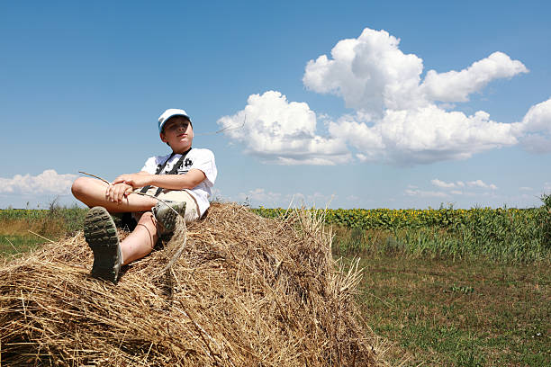 Boy resting on haystack in rural field with blue sky. Child enjoying summer day outdoors in nature. Countryside peaceful scene.