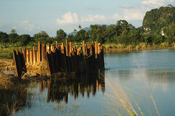 Steel sheet piles driven into the ground beside a calm river, surrounded by lush greenery and distant hills, illustrating civil engineering in a natural setting.