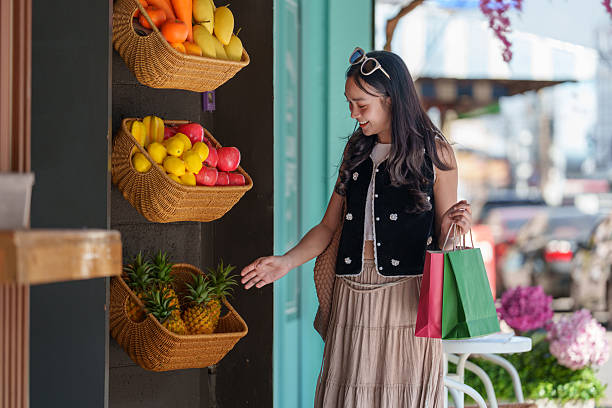 Young woman smiling while selecting fresh pineapples from wicker baskets at a vibrant greengrocer's stall, holding shopping bags and enjoying a healthy lifestyle in the summer sun
