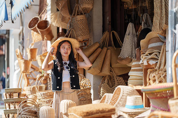 istock Young woman tourist wearing a straw hat adjusts her hat while browsing wicker bags, baskets, and other handmade crafts at a traditional market stall 2253080103