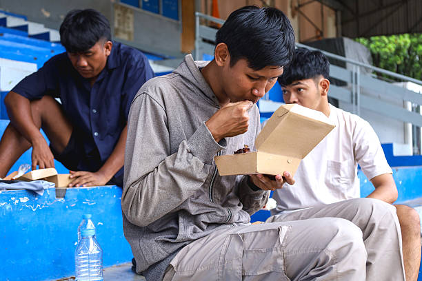 Young Men Sharing a Meal Together in Outdoor Stadium Seating, Enjoying Each Other's Company in a Relaxed Atmosphere