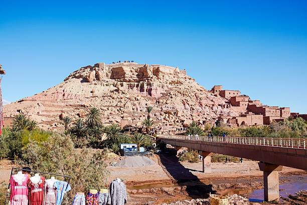 Morocco, Ait Benhaddou, November 16, 2025: View of the modern pedestrian bridge crossing the shallow river towards the fortified hilltop ksar, with palm trees along the banks and colorful traditional dresses displayed on mannequins in the foreground under a bright blue sky.