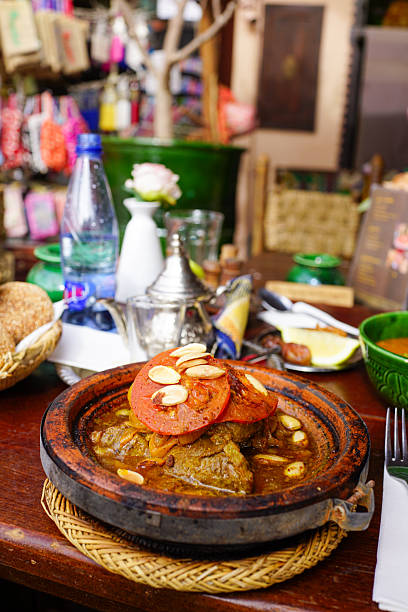 Morocco, Ait Benhaddou, November 16, 2025: Close-up of a traditional meat tajine served in an earthenware dish, topped with tomato slices and almonds on a wooden table, with bread, mint tea pot, water bottle and colorful decor of a cozy restaurant softly blurred in the background.