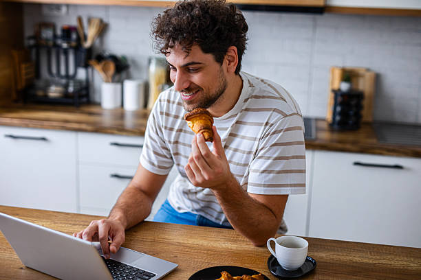 istock A smiling man eats a croissant while working on his laptop at home, representing a relaxed and productive remote work lifestyle 2253097393