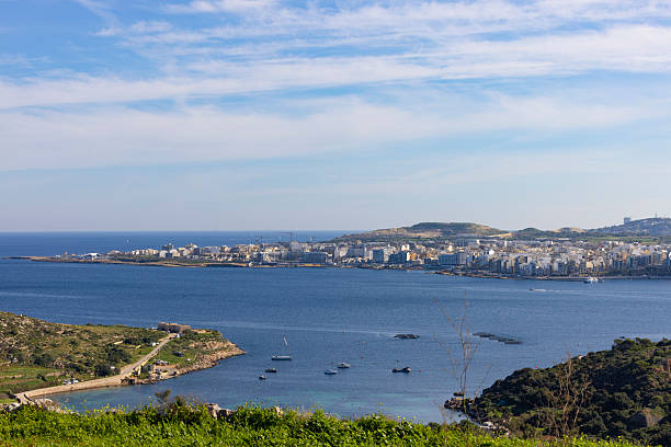 View over a coastal bay in Malta with calm blue Mediterranean water, small boats anchored in the bay, and a distant city skyline along the coastline under a bright sky.
