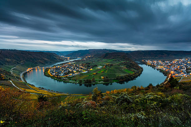 Moselle River meander in western Germany, with illuminated villages tracing the looping river bend through the valley. Farmland and vineyards lie at the center of the curve, while dark storm clouds hang overhead, creating a dramatic and atmospheric scene in the Moselle Valley, Rhineland-Palatinate, Germany - Europe