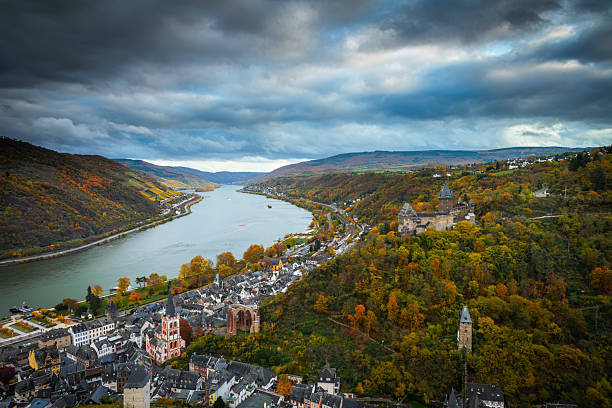 Historic town of Bacharach along the Rhine River in western Germany, surrounded by forested hills with vibrant autumn foliage. The scenic river valley features medieval architecture, church towers, vineyards, and dramatic clouded skies, highlighting the Upper Middle Rhine Valley, a UNESCO World Heritage site and popular European travel destination. Bacharach in Rhineland-Palatinate, Germany - Europe