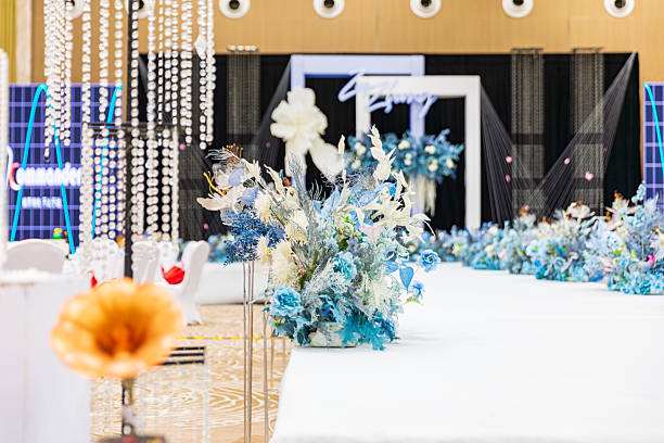 A close-up of clear tall candle holders and lush floral arrangements in yellow and white that run along the table. In the blurred background are round banquet tables with white chair covers and gift bags, warm lighting producing a soft glow. The shallow depth of field emphasizes the texture and colors of the centerpiece while conveying an elegant, festive mood.