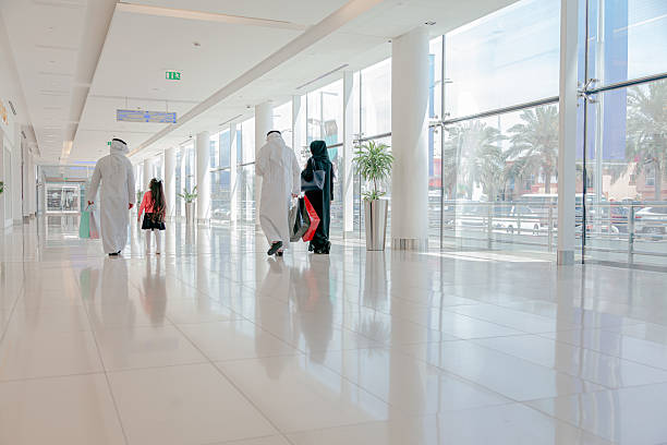 A Middle Eastern family including two adult men, one adult woman and a young child walking together in a spacious modern indoor corridor with floor to ceiling windows, reflective marble floor and natural daylight, wearing traditional Gulf clothing and abaya, representing family lifestyle, togetherness, leisure time and contemporary Middle Eastern culture in a clean architectural interior with strong copy space