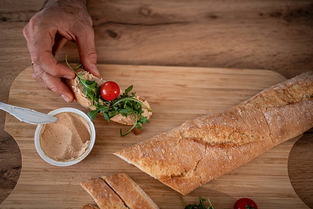 Person's hand making a small sandwich with pate spread on a slice of fresh baguette, topped with arugula and a cherry tomato on a wooden cutting board