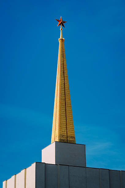 Golden obelisk tower rises above a white building against a clear blue sky, adorned with a red star at the tip. A historic Leipzig landmark captured in bright daylight.soviet Pavilion on the former trade fair grounds, later known as the Achilleion